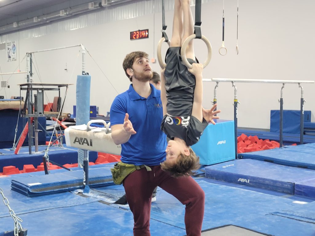 A boy doing a movement on rings with assistance of coach