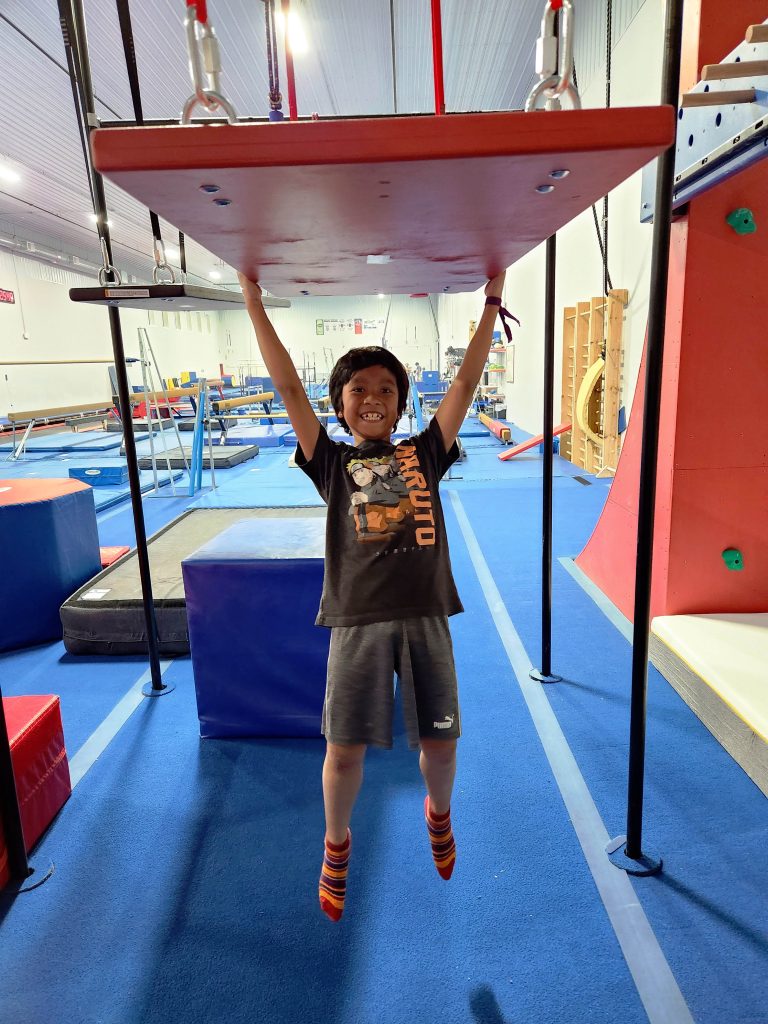A boy hangs from a suspended board as part Ninja Parkour class