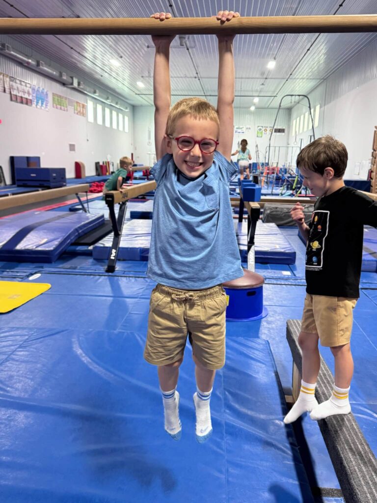 Camper smiling while hanging from the gymnastics bars during Camp ROGA's Olympic-themed summer camp week.